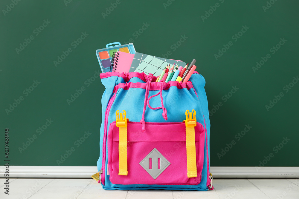 Colorful school backpack with notebooks, watercolors and pencils on white tile table near green chal