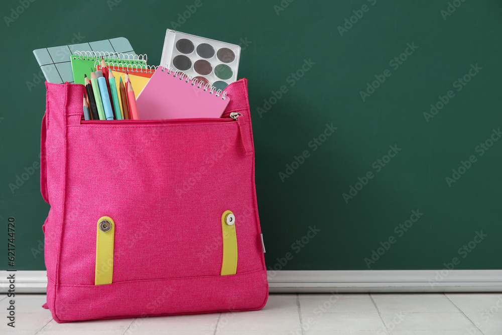 Pink school backpack with notebooks, watercolors and pencils on white tile table near green chalkboa