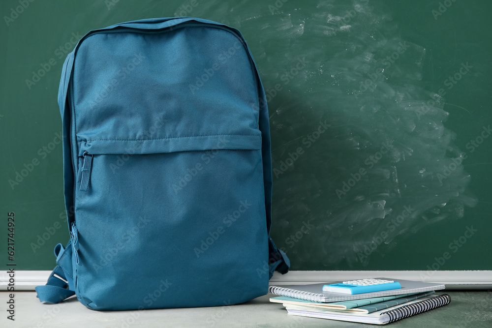 Blue school backpack with notebooks and calculator on table near green chalkboard