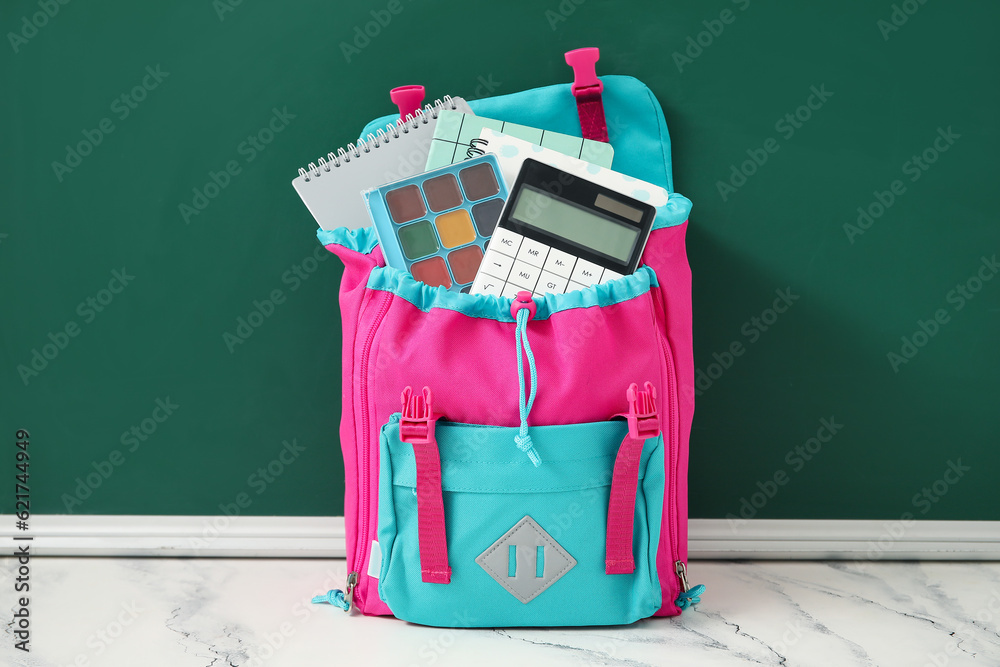 Colorful school backpack with notebooks, watercolors and calculator on white table near green chalkb