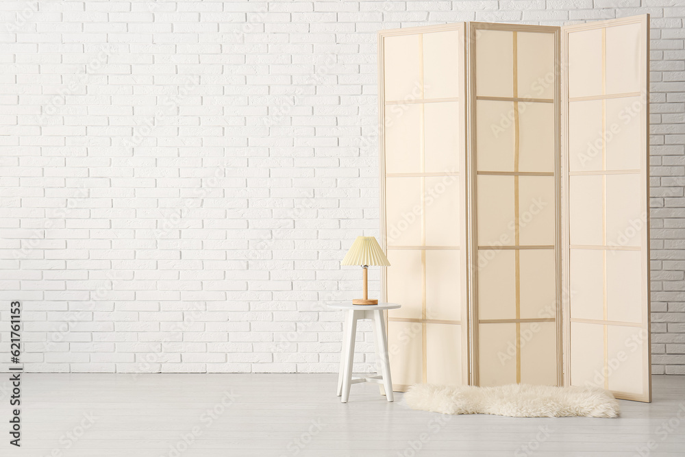 Folding screen with chair and lamp near white brick wall