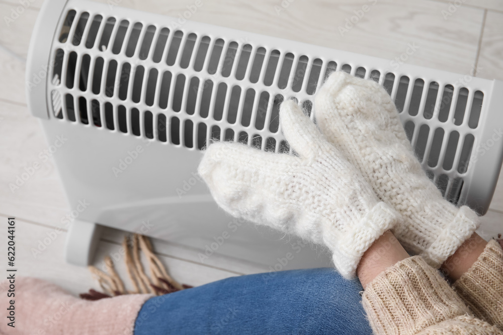 Woman warming hands in mittens near radiator at home, closeup