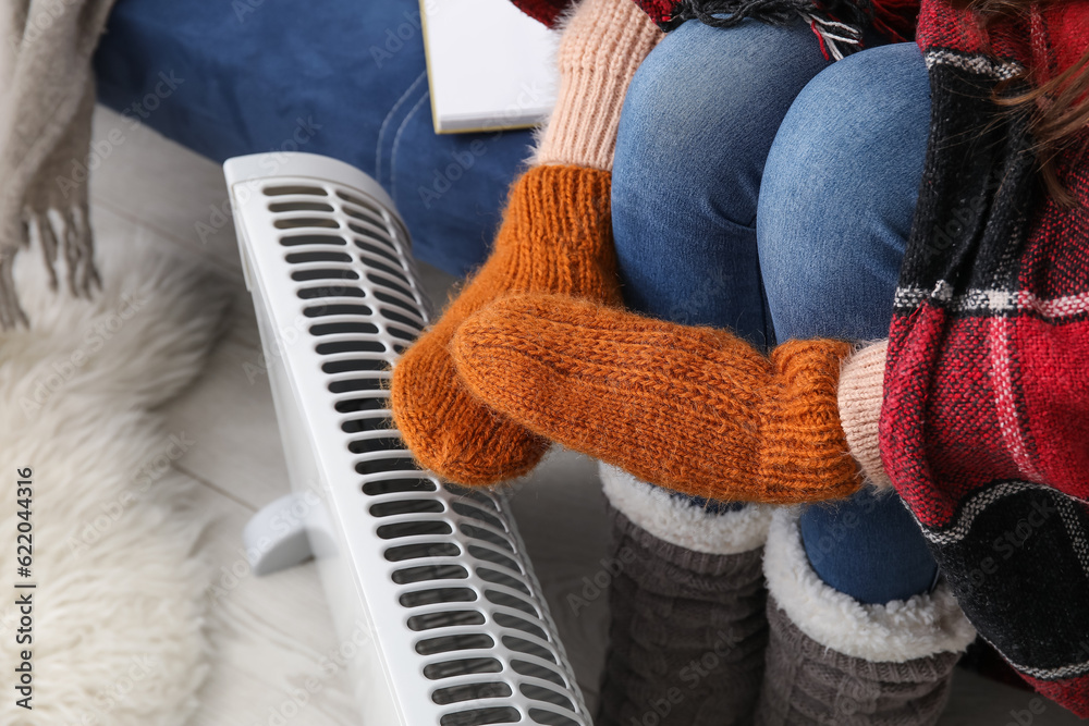 Woman warming hands in mittens near radiator at home, closeup