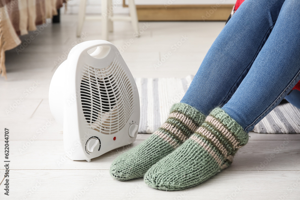 Woman warming legs in socks near electric fan heater at home, closeup