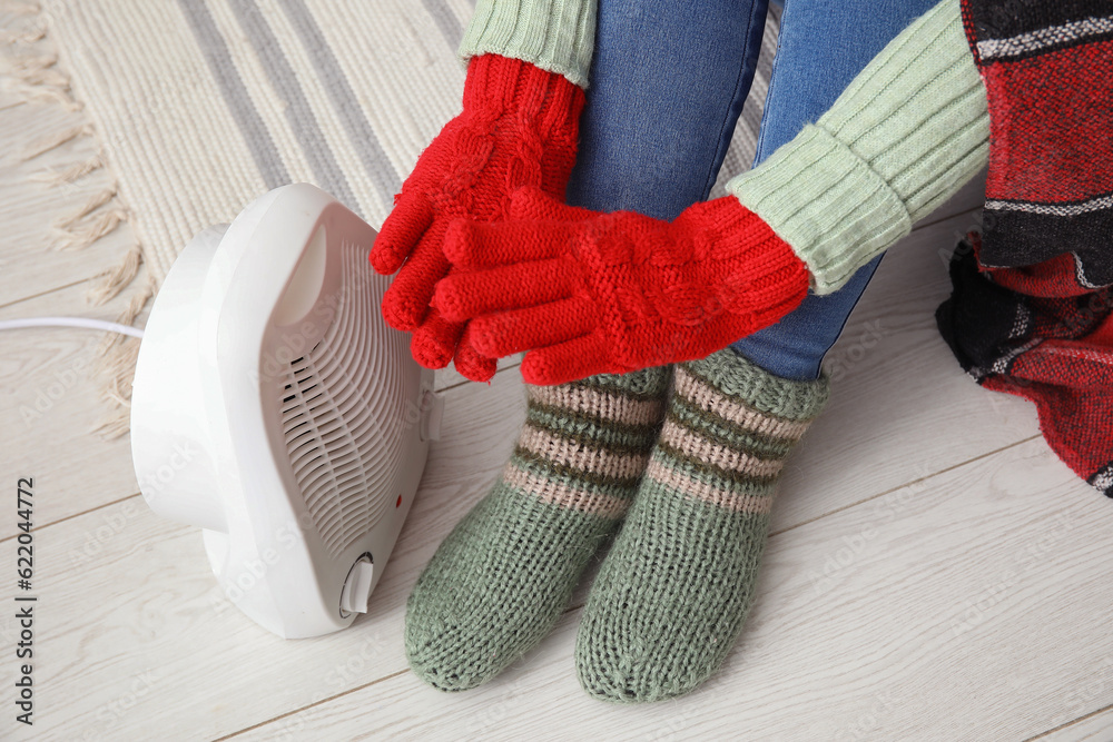 Woman warming hands in gloves near electric fan heater at home, closeup