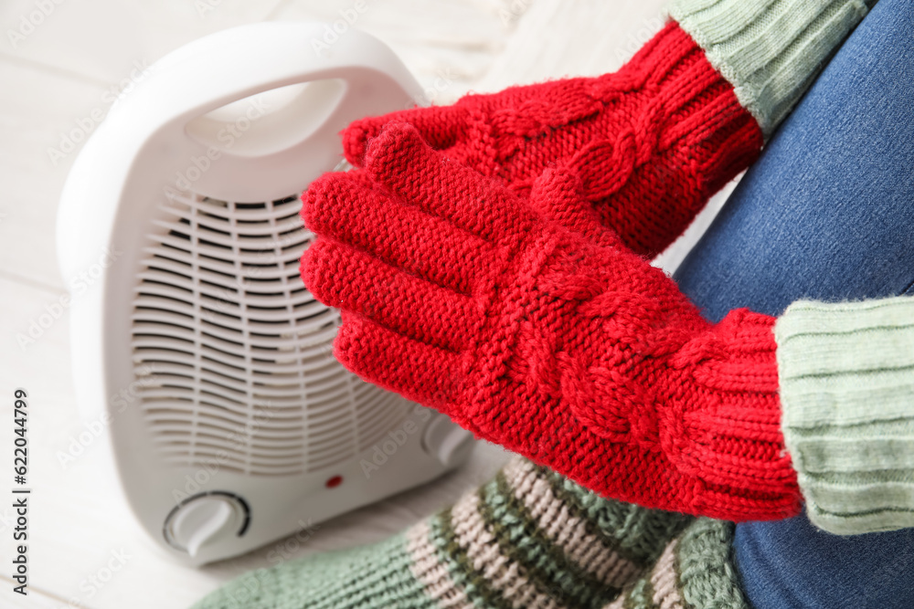 Woman warming hands in gloves near electric fan heater at home, closeup