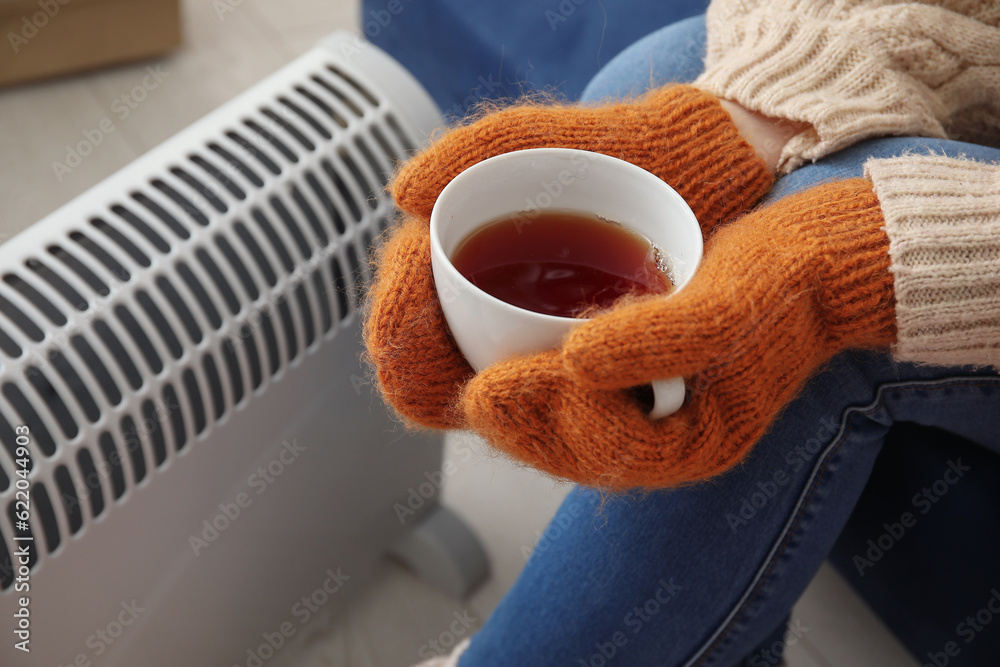 Woman with cup of hot coffee warming near radiator at home, closeup