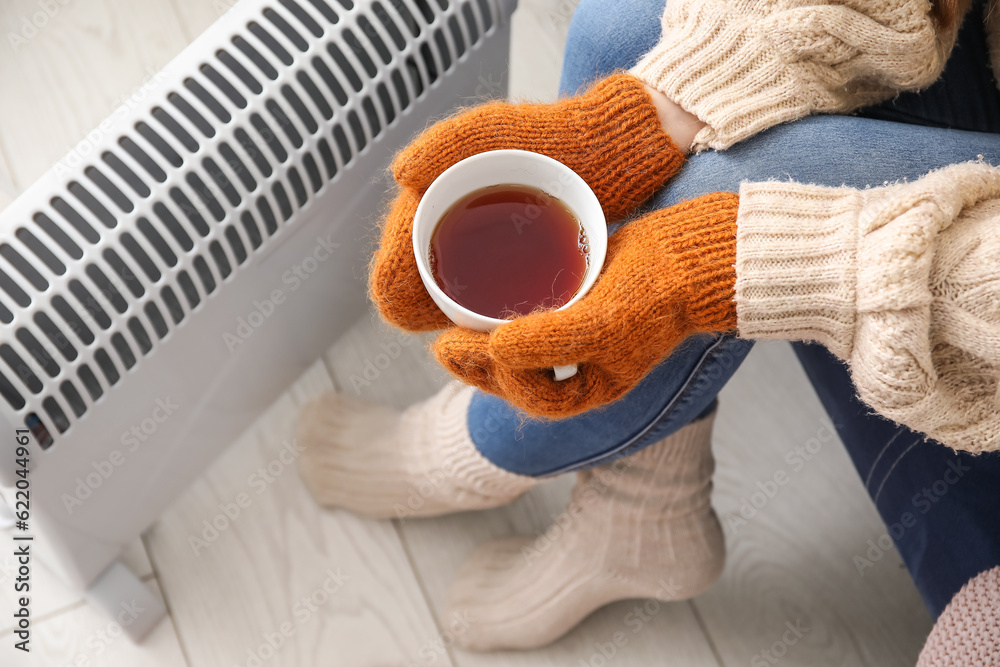 Woman with cup of hot coffee warming near radiator at home, closeup