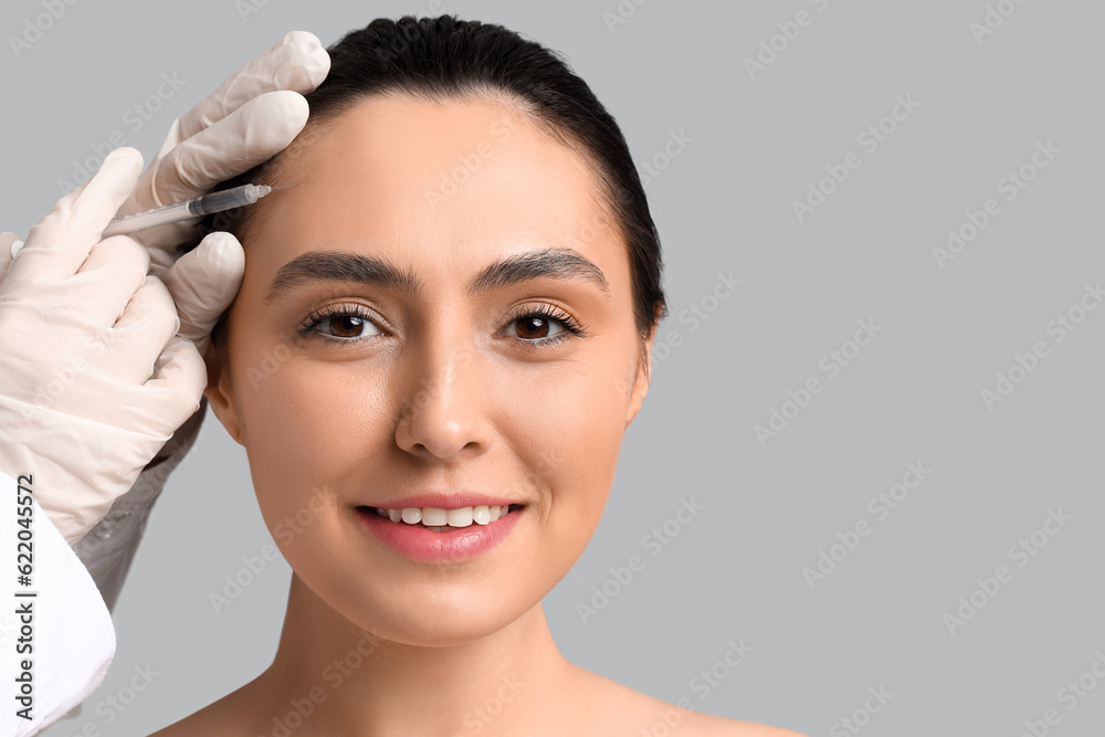 Young woman receiving filler injection in face against grey background