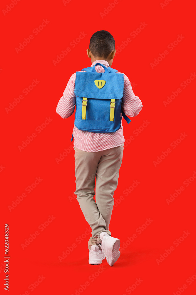 Little African-American schoolboy with backpack on red background, back view