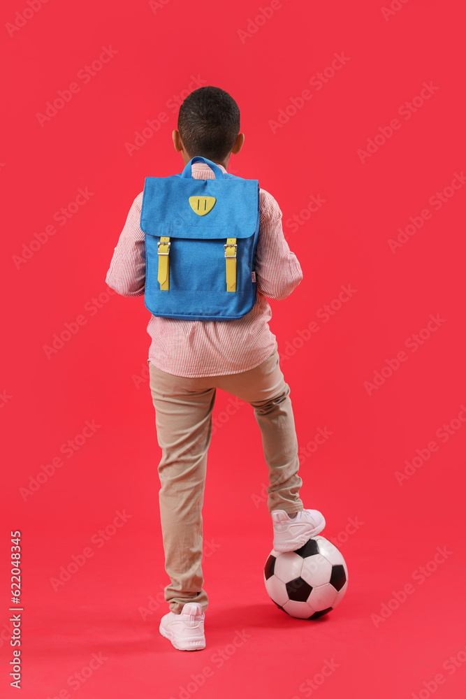 Little African-American schoolboy with soccer ball and backpack on red background, back view