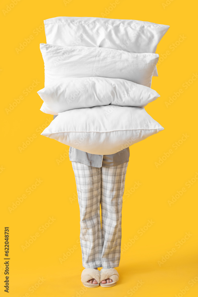 Young woman in pajamas and with pillows on yellow background