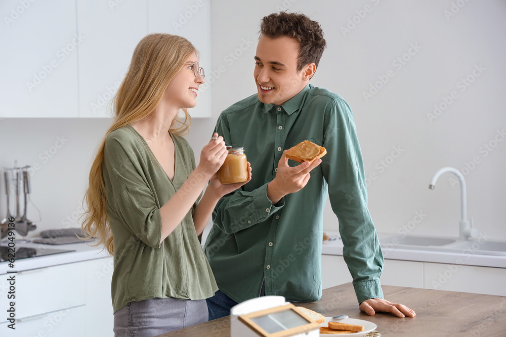 Young woman holding jar of nut butter and handsome man with toast in kitchen