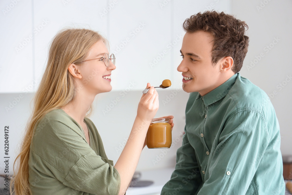 Young woman feeding her boyfriend with tasty nut butter in kitchen