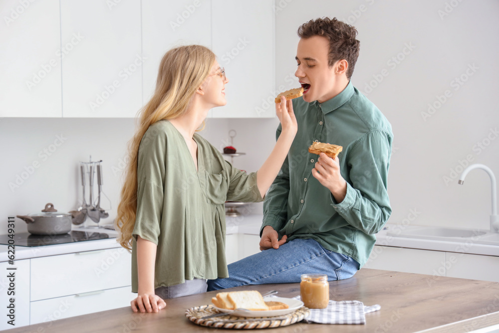 Young woman feeding her boyfriend with tasty toast of nut butter in kitchen