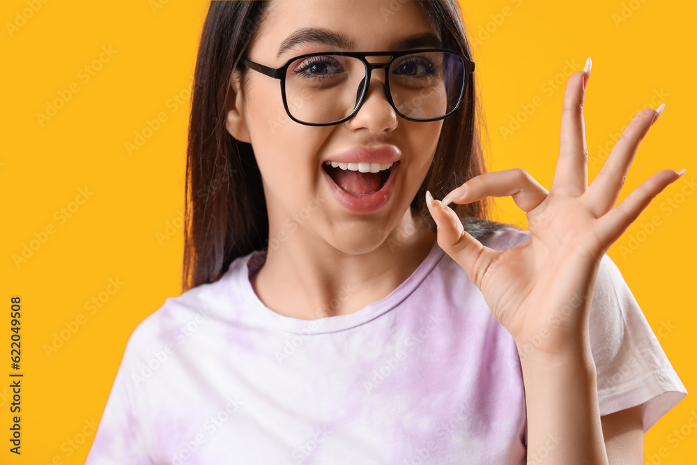 Pretty young woman wearing glasses and showing OK on yellow background