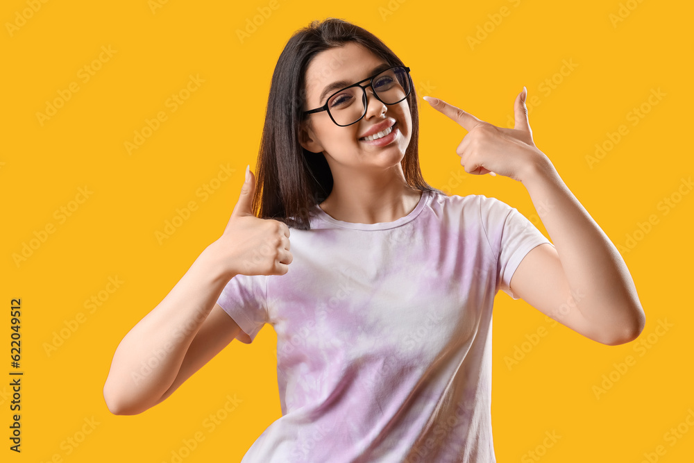 Pretty young woman wearing glasses and showing thumb-up on yellow background