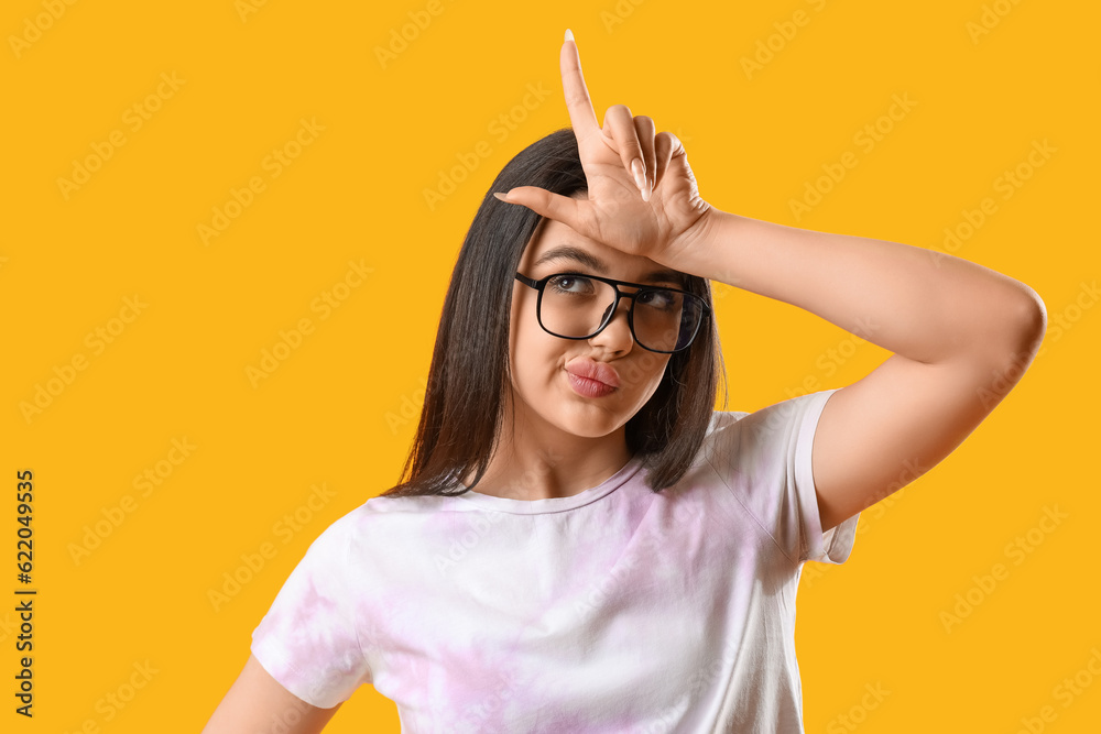 Pretty young woman wearing glasses and showing  loser  gesture on yellow background