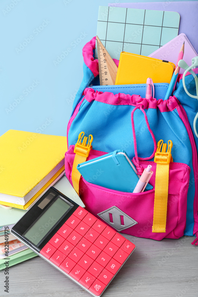 Color school backpack with calculator, books and eyeglasses on grey wooden table near blue wall