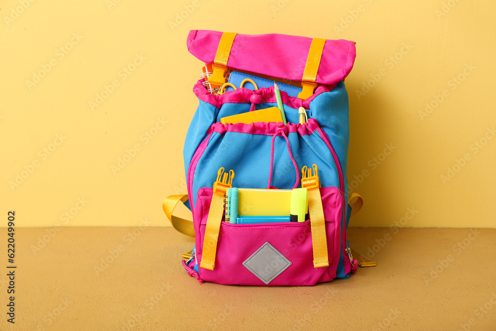 Color school backpack with notebooks, pens and marker on table near yellow wall