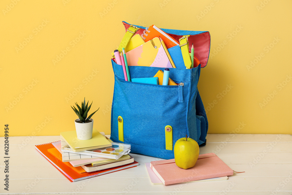 Blue school backpack with notebooks, houseplant and apple on white wooden table near blue wall