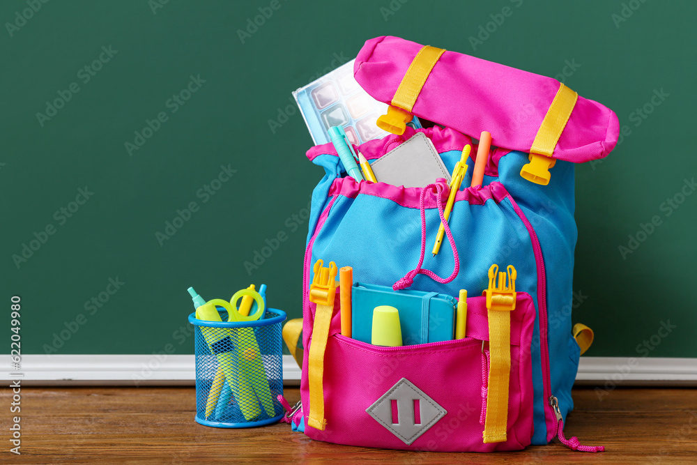 Color school backpack with markers, pens and watercolor on brown wooden table near chalkboard