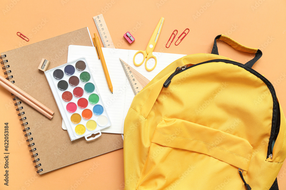 School backpack with paints and stationery on orange background