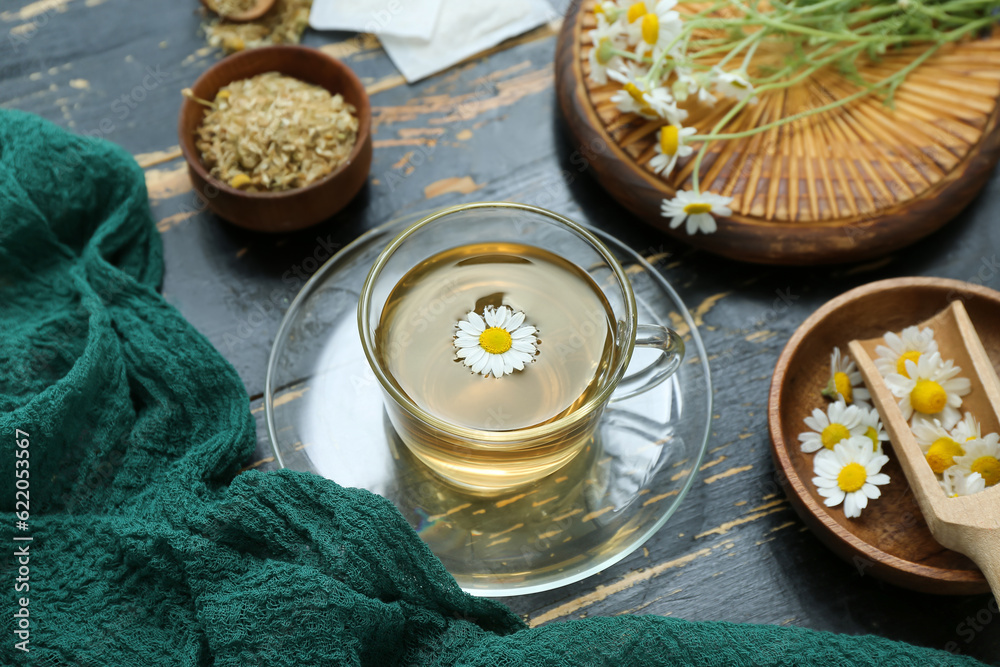 Cup of hot tea and bowl with dried chamomile on black wooden background