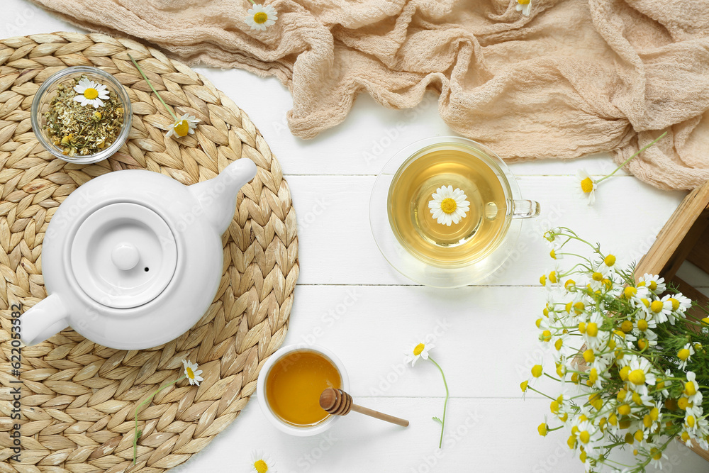 Wicker mat with honey, teapot and cup of hot chamomile tea on white wooden background