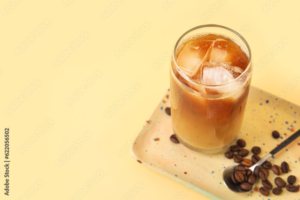 Glass of ice coffee with beans, spoon and plate on yellow background