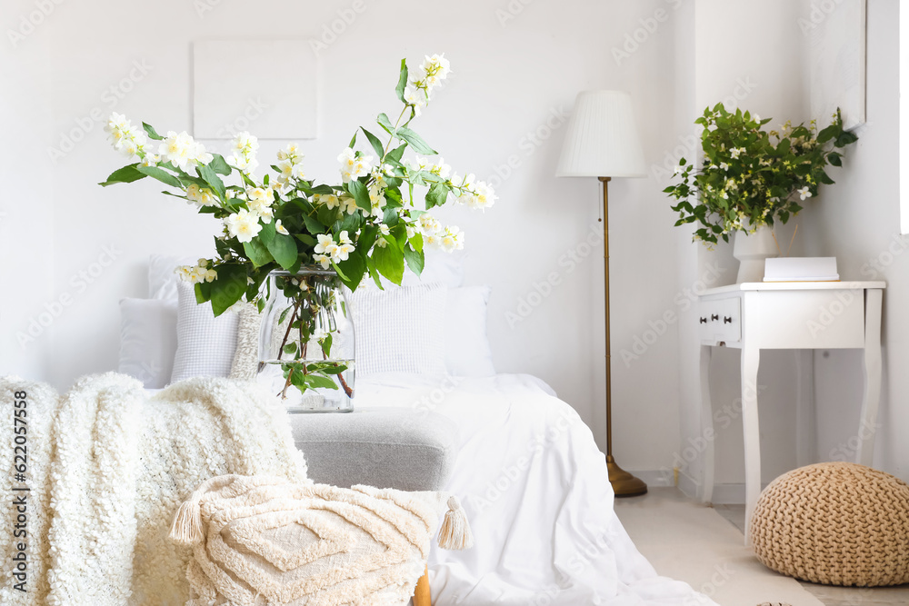 Vase with blooming jasmine flowers on bedside bench in interior of light bedroom