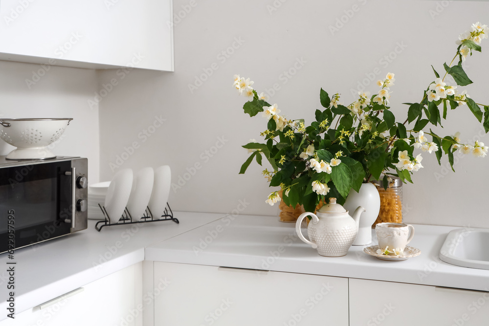 Vase with blooming jasmine flowers, teapot and cup of tea on white kitchen counter