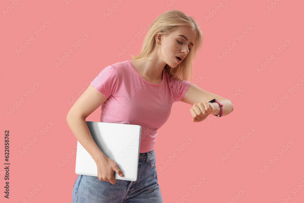 Stressed young woman with laptop looking at wristwatch on pink background. Deadline concept