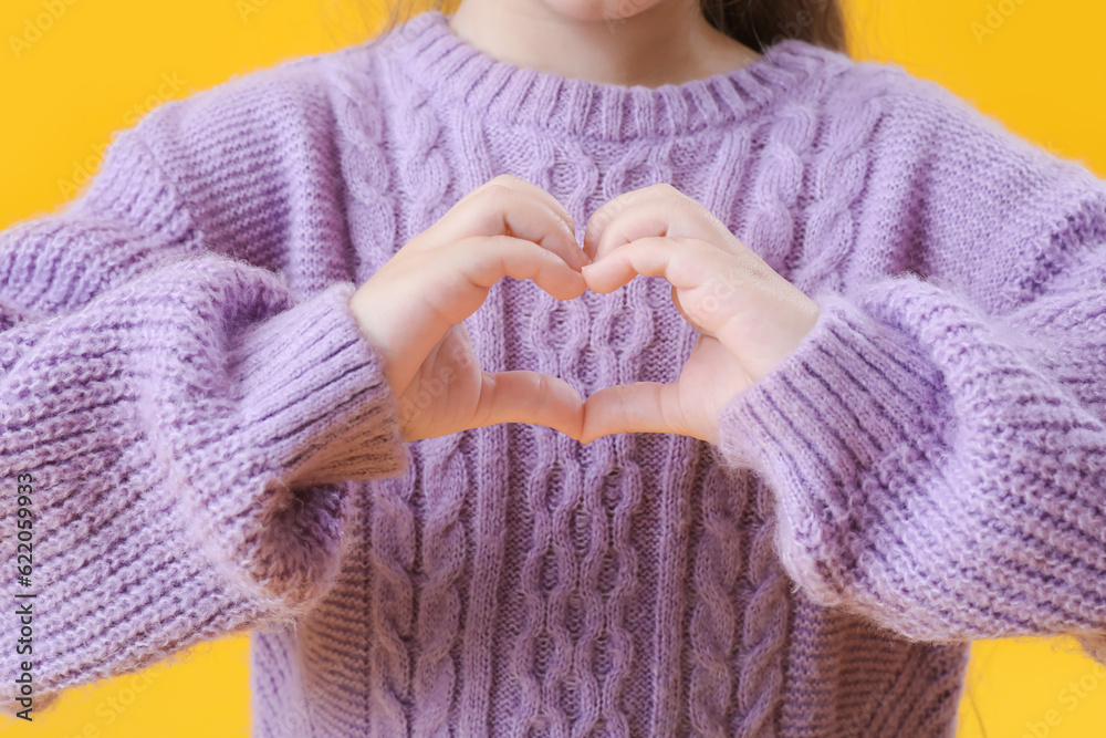 Cute little girl in knitted sweater making heart with her hands on yellow background, closeup