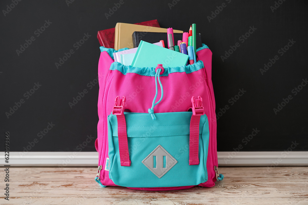 Pink school backpack with notebook and pencils on wooden table near black chalkboard