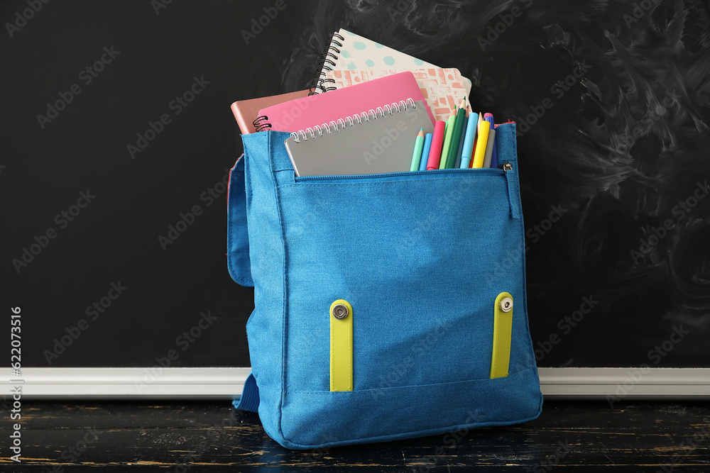 Blue school backpack with notebooks, pencils and markers on wooden table near black chalkboard