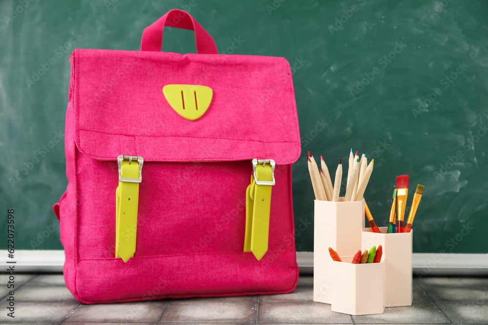 Pink school backpack with cup of pencils and brushes on grey tile table near green chalkboard