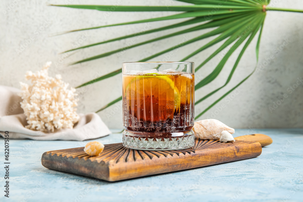 Board with glass of cold Cuba Libre cocktail and seashells on blue table