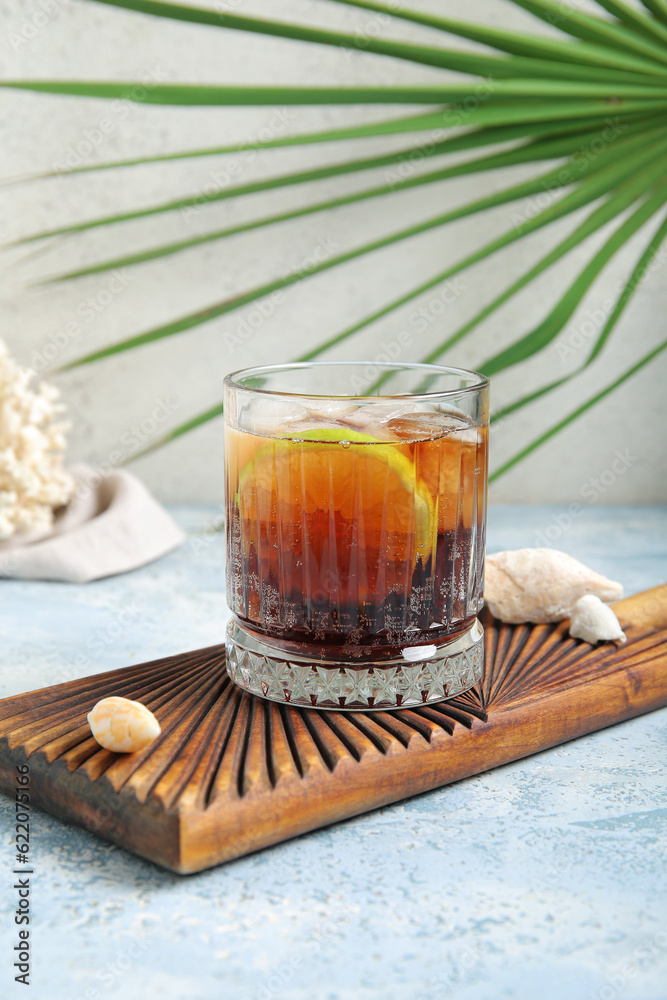 Board with glass of cold Cuba Libre cocktail and seashells on blue table