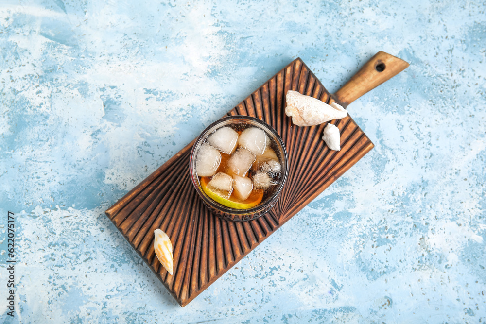 Board with glass of cold Cuba Libre cocktail and seashells on blue background