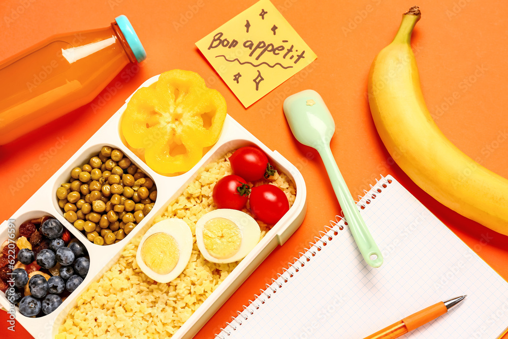 Stationery, tasty food in lunchbox and sticky note with text BON APPETIT on orange background