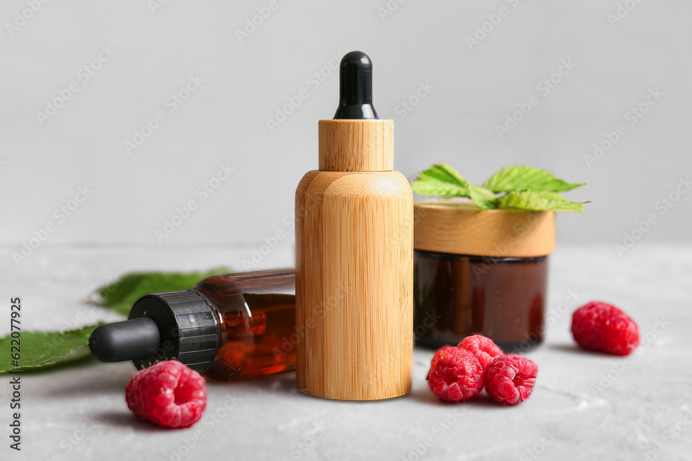 Bottles of cosmetic raspberry oil and jar with cream on grey table