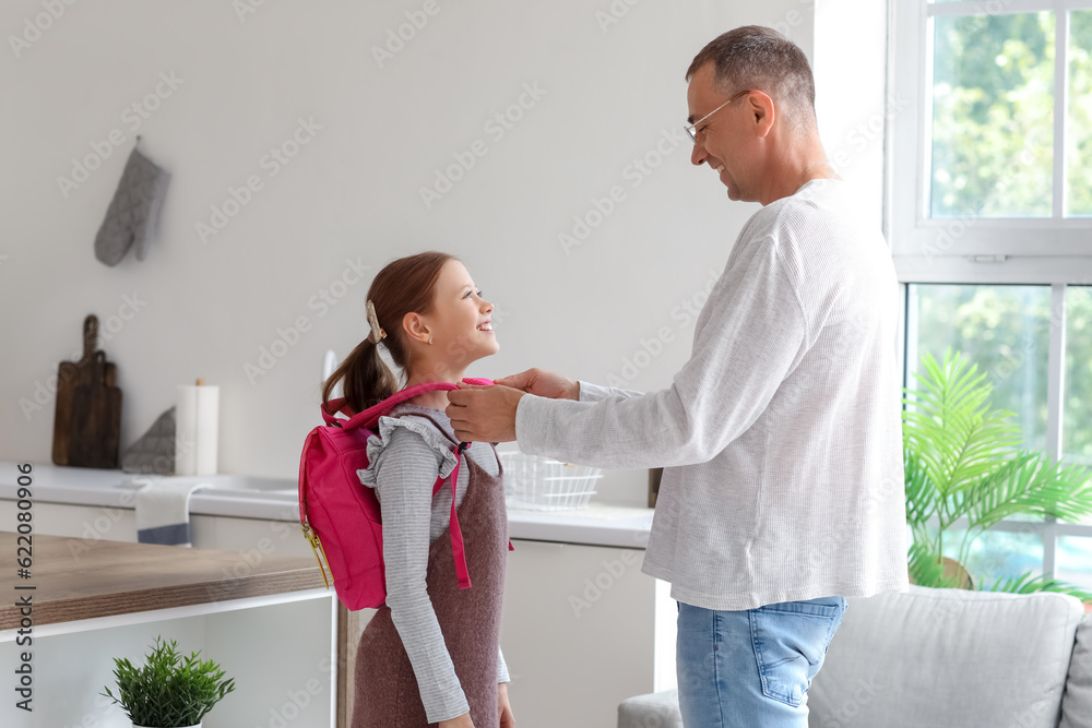 Father helping his little daughter to put schoolbag on in kitchen