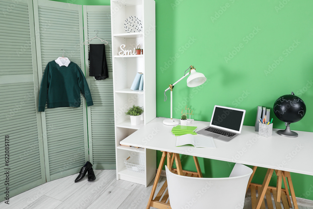 Interior of workplace with desk, shelving unit, laptop computer, shoes and stylish school uniform ha