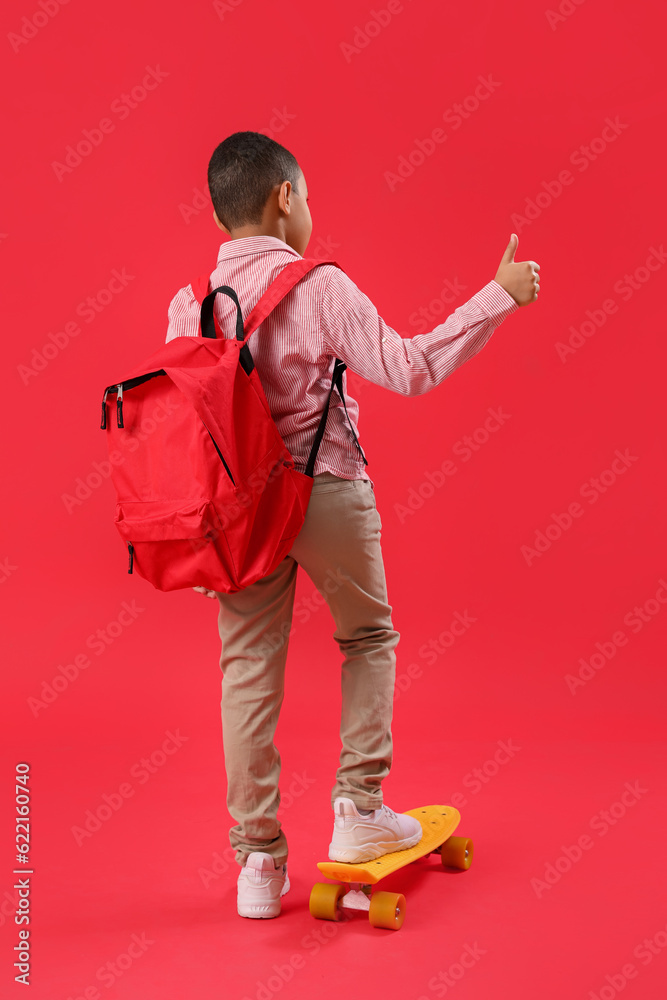 Little African-American schoolboy with backpack and skateboard showing thumb-up on red background, b