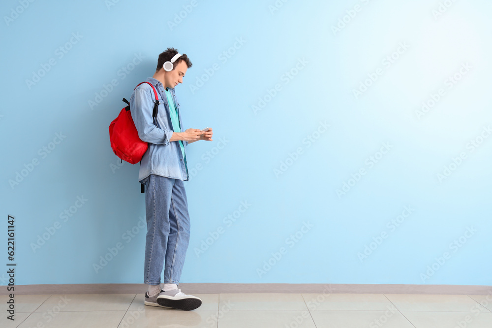 Male student with backpack and mobile phone listening music in headphones near blue wall