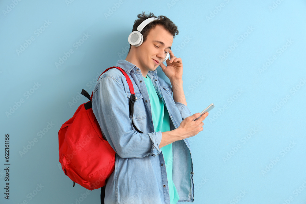 Male student with backpack and mobile phone listening music in headphones on blue background