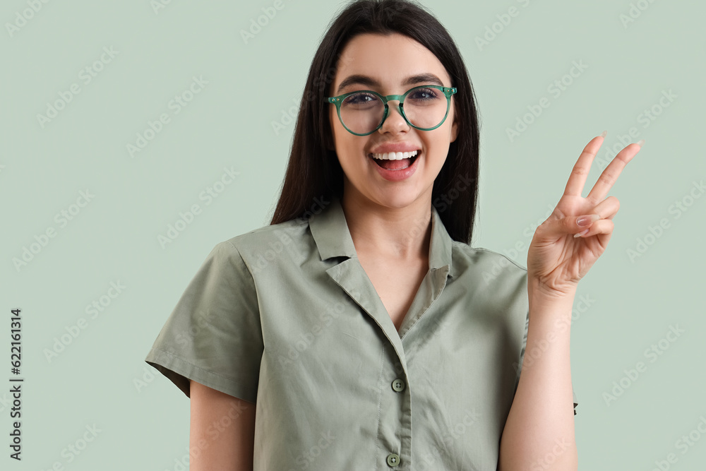 Beautiful young woman wearing glasses and showing victory gesture on color background