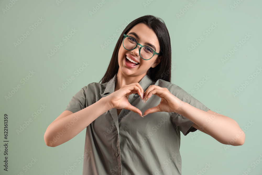 Beautiful young woman wearing glasses and making heart with her hands on color background