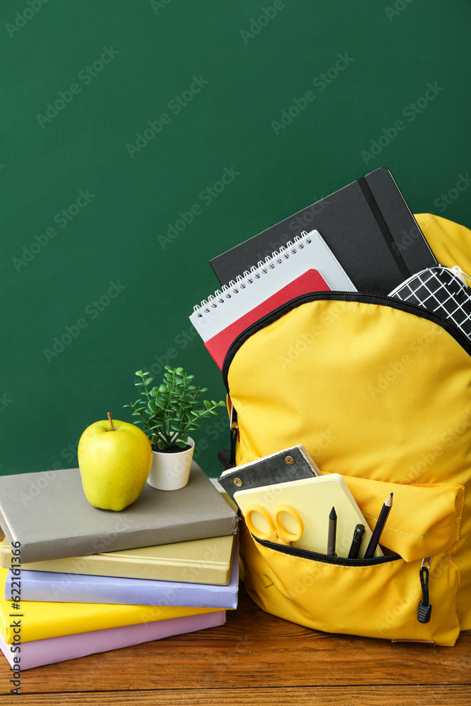 Yellow school backpack with books, houseplant and apple on wooden table near chalkboard